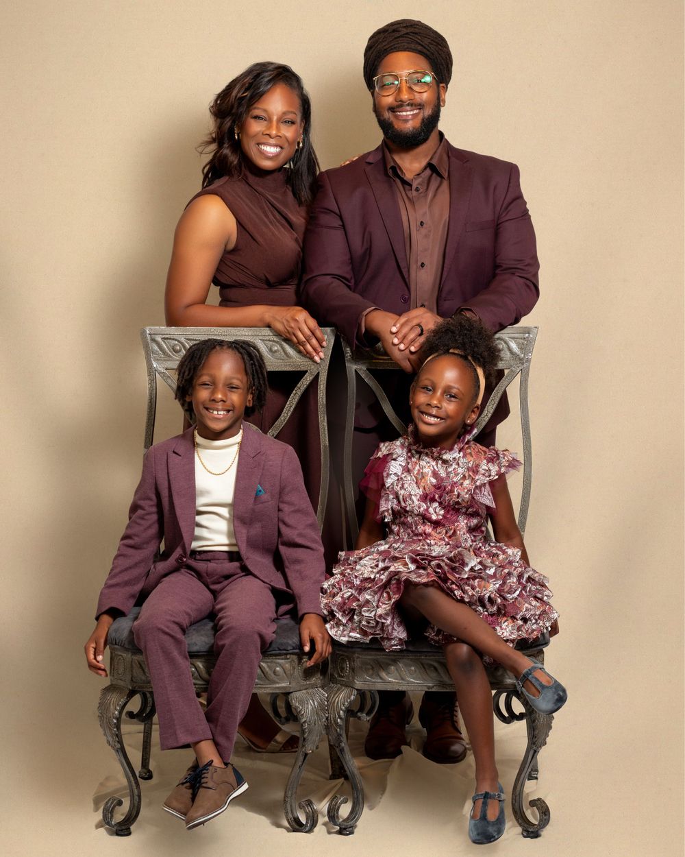 A stylish family of four wearing coordinated burgundy outfits poses together on elegant vintage chairs against a neutral backdrop.