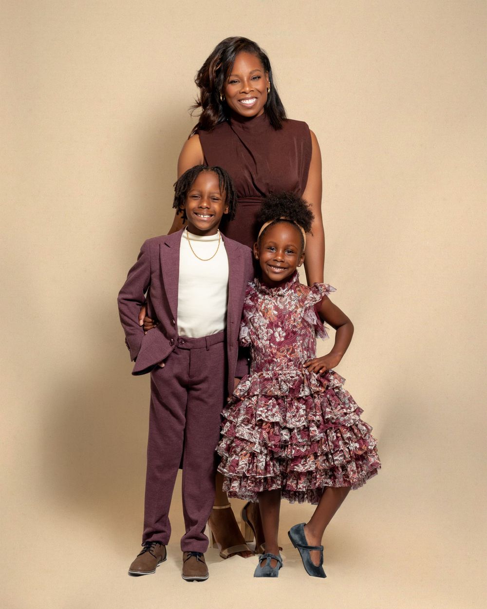 Three people pose together against beige backdrop wearing coordinated burgundy and floral outfits for a formal portrait.