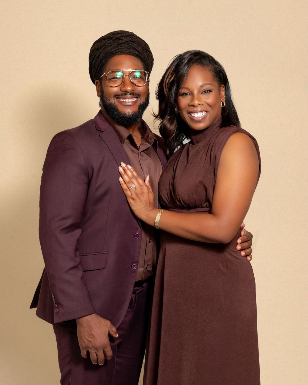 A couple in matching burgundy formal attire pose intimately together for a portrait against a beige background.