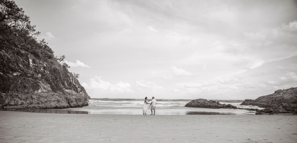 Two silhouettes stand together on a scenic black and white beach surrounded by rocky cliffs and ocean waves.