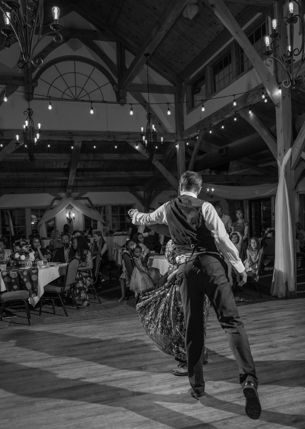 A dramatic black and white shot of dancers performing in a barn-style venue with string lights.
