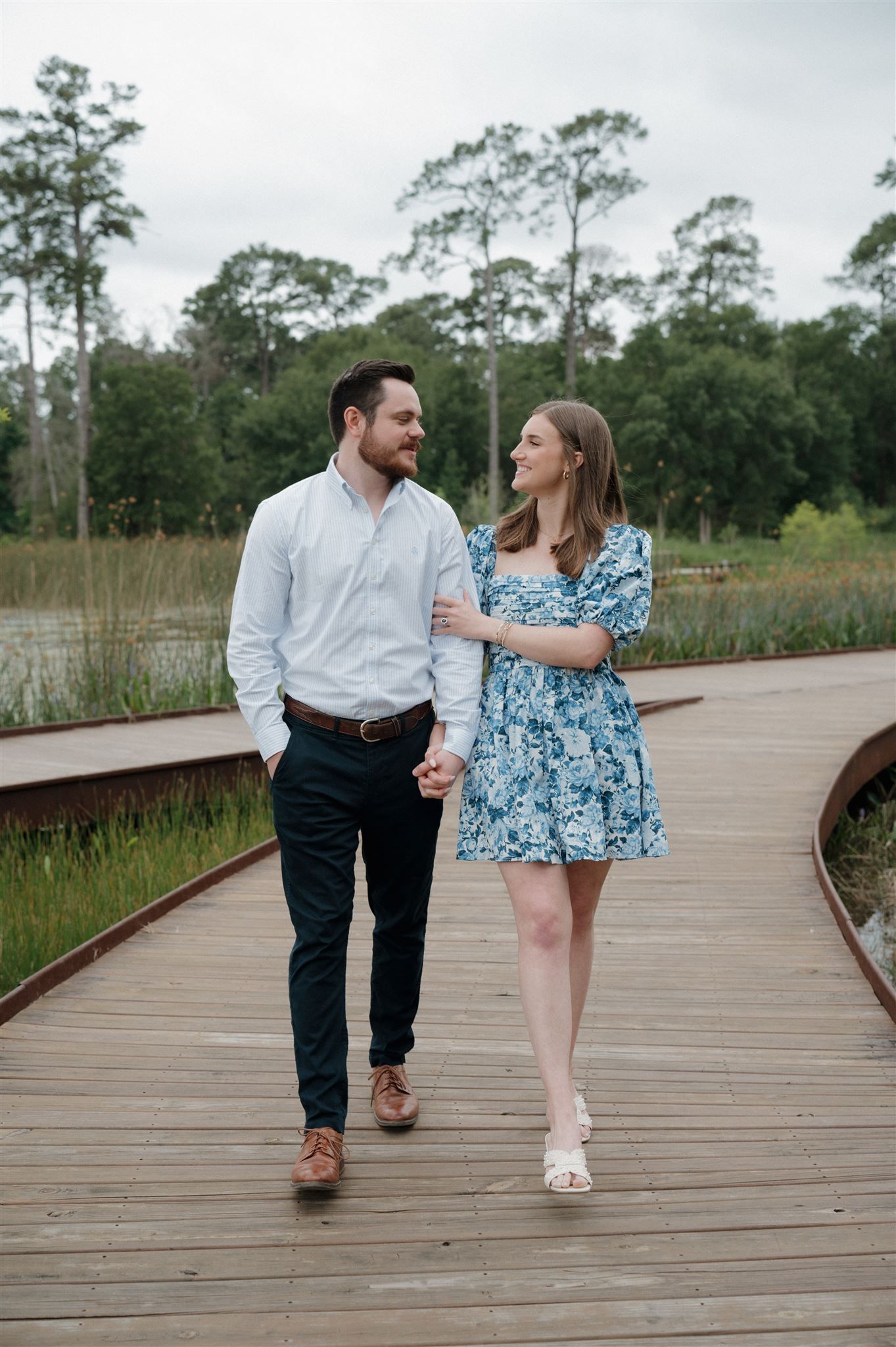 A couple takes a romantic stroll along a wooden boardwalk surrounded by tall pine trees and marsh grasses.