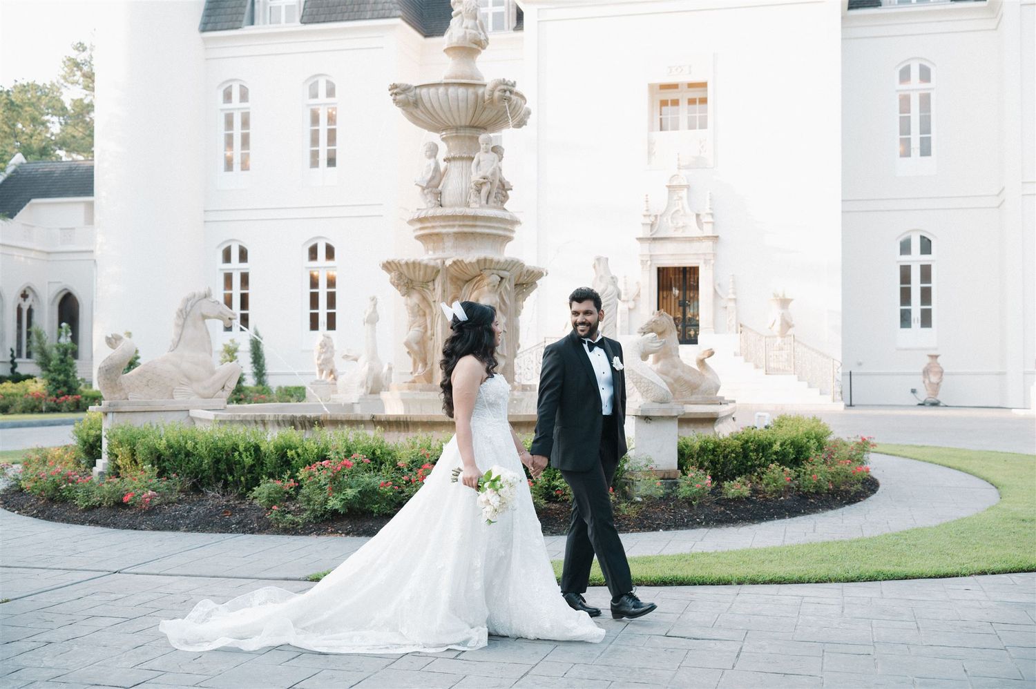 Elegant wedding couple walks past ornate fountain in front of grand white mansion with classical architecture.
