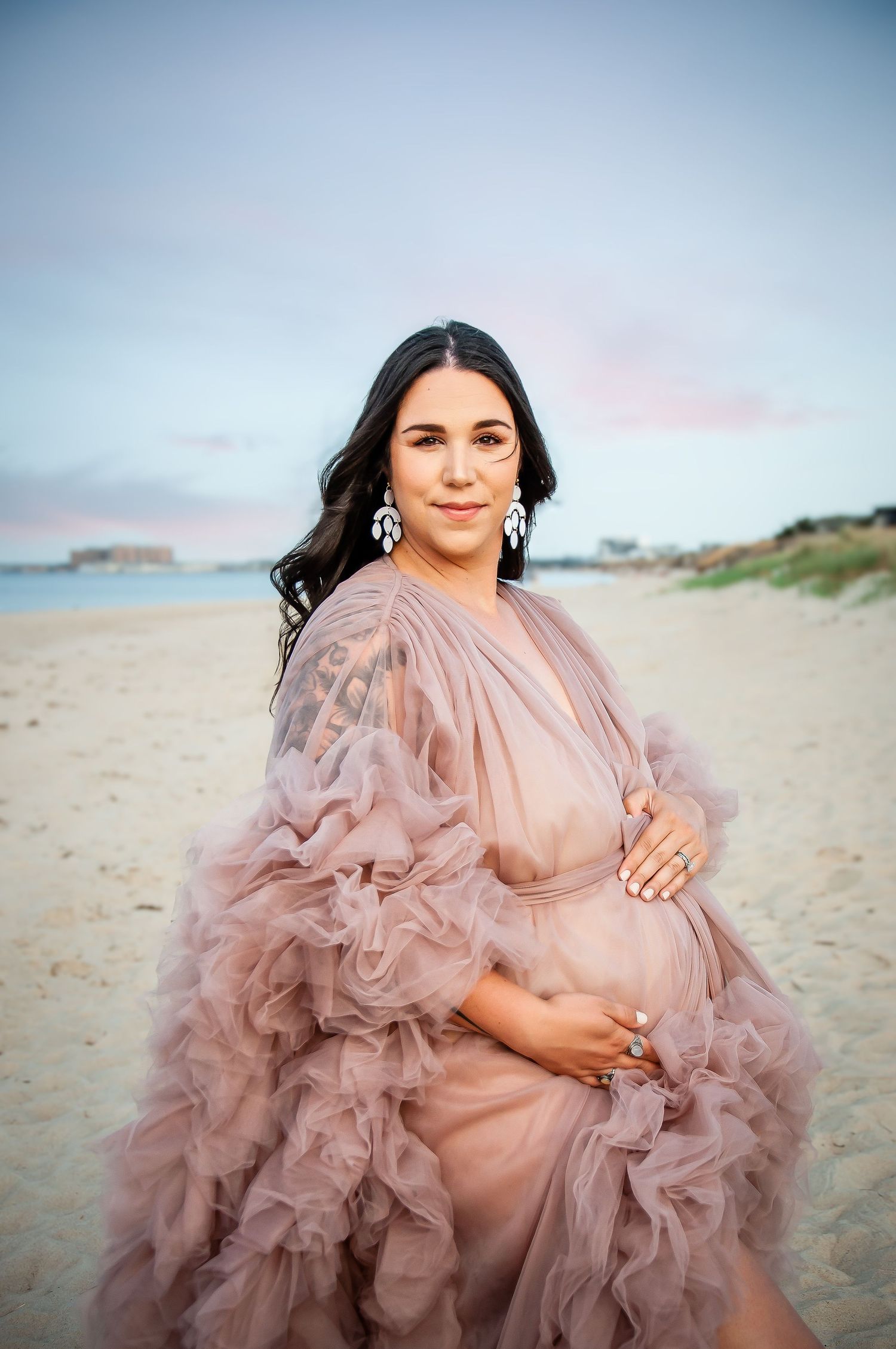 Pregnant person in flowing pink tulle gown poses on beach at sunset.