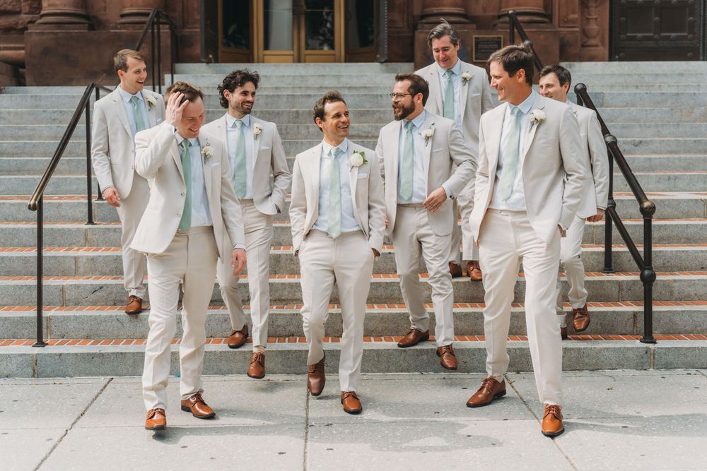 Groomsmen in light colored suits pose together on stone steps showing off their brown dress shoes.