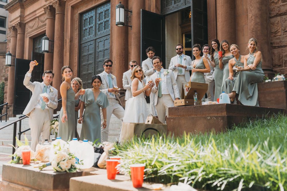 Wedding party in white and light colored attire posing on church steps outdoors.