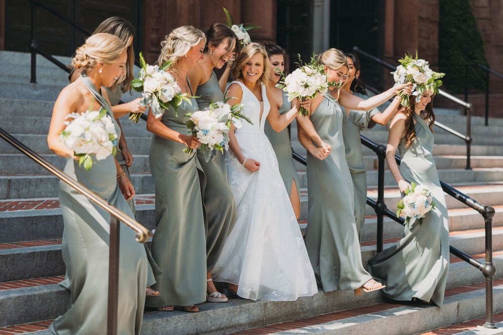 Wedding party in sage green dresses and white gown pose together on stone steps.