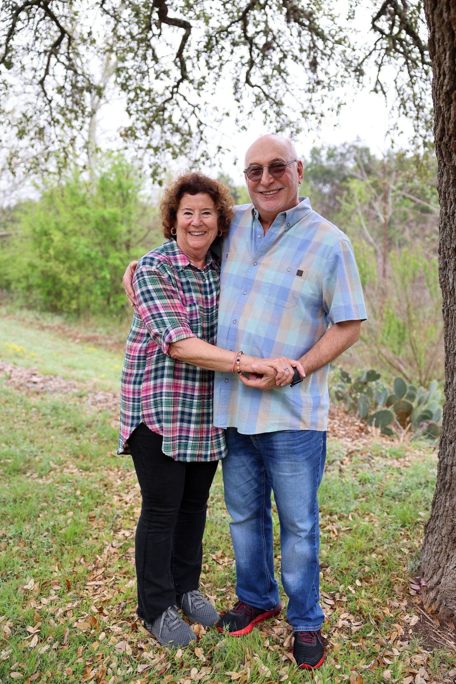 A couple embracing outdoors under a tree in casual clothing.