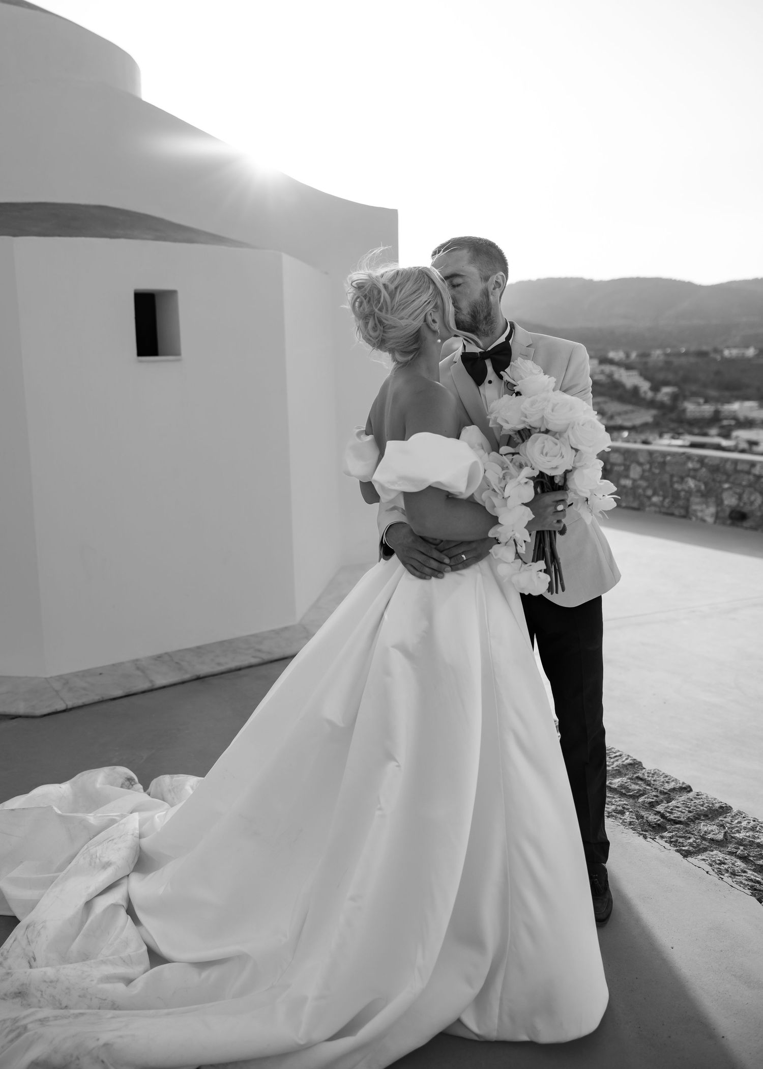 A romantic black and white wedding photo captures a couple embracing on a greek terrace with a scenic mountain view at a wedding.