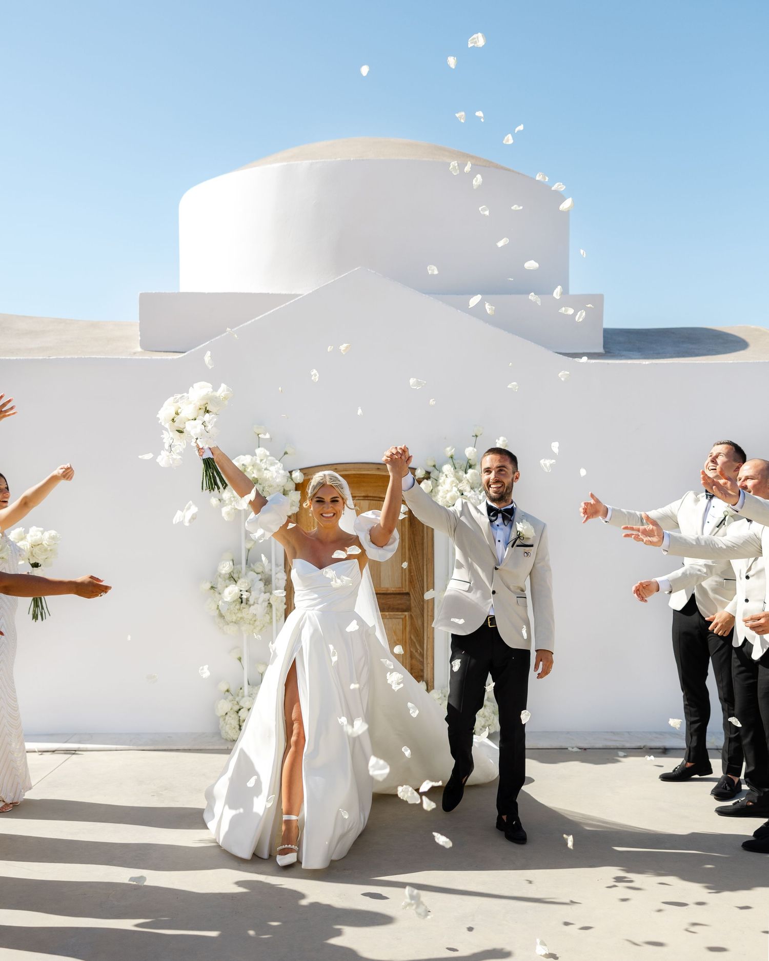 A joyful wedding party celebrates with white flower petals being tossed in front of a modern white building in Lindos, Greece.
