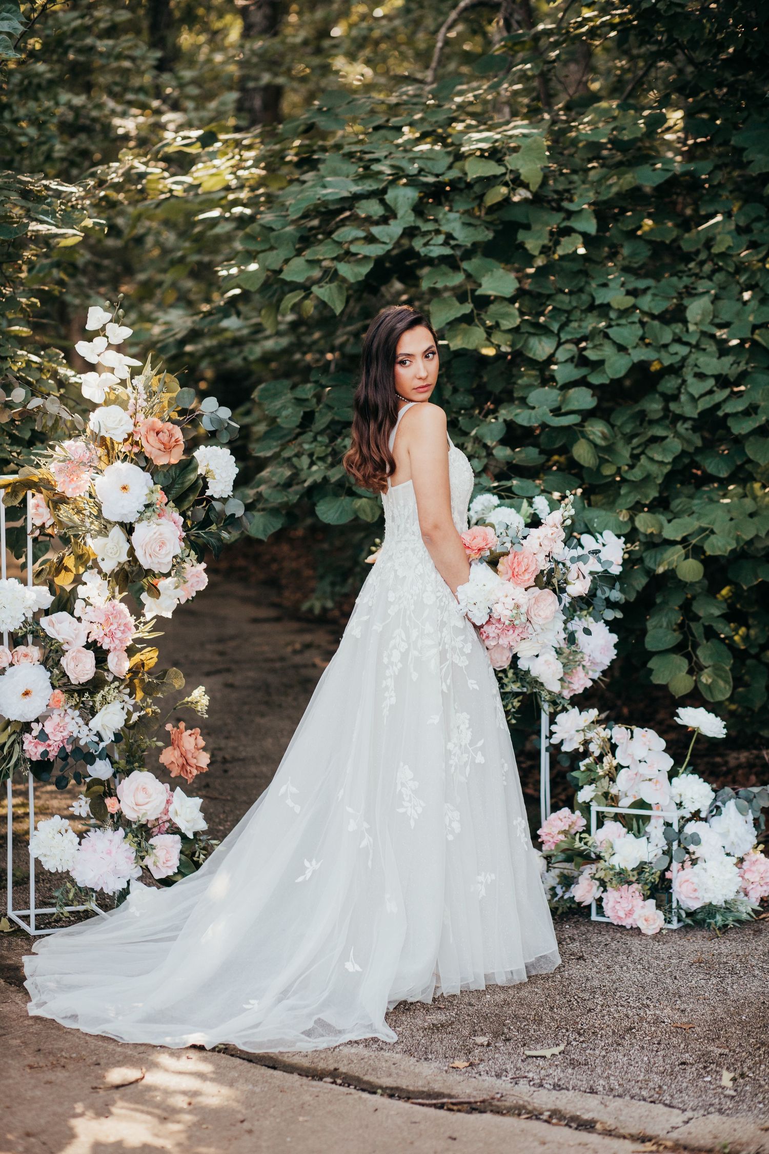 A bride in a flowing white lace gown poses next to an elaborate floral arch with pink and white blooms in a garden setting.