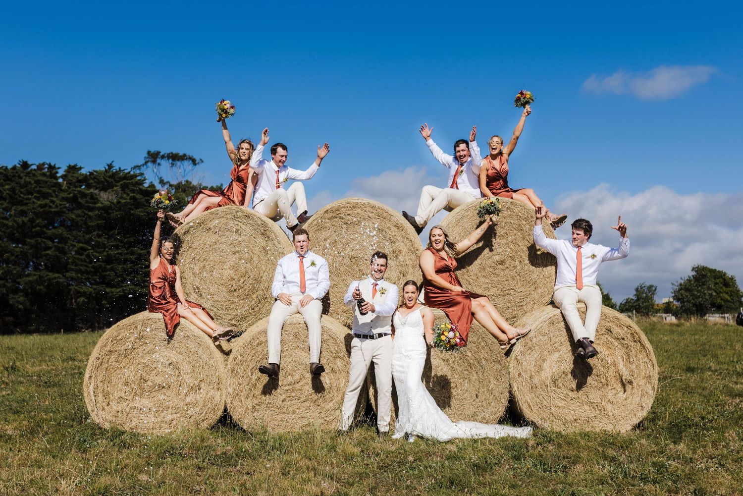 Wedding party celebrating on hay bales in a sunny field with joyful poses.