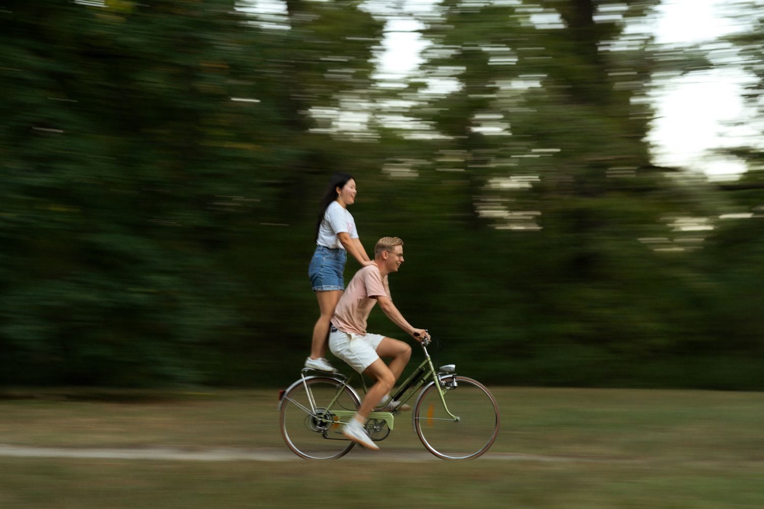A motion-blurred shot of two people riding together on a vintage bicycle through a green wooded area.