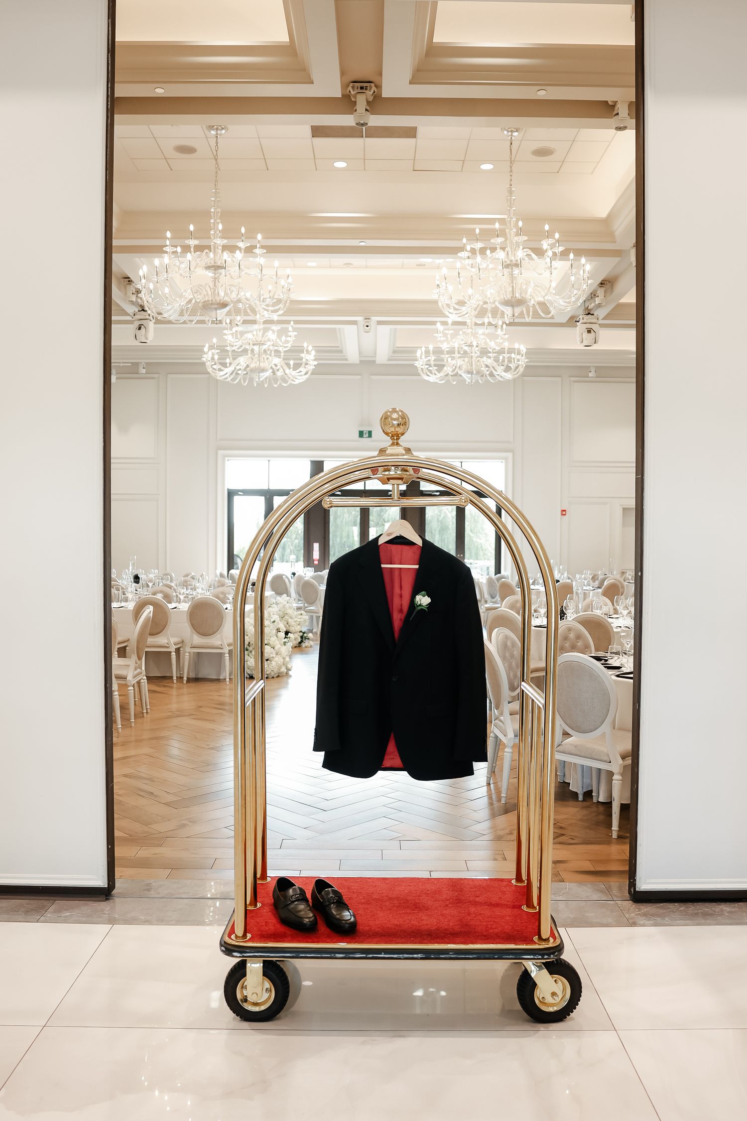 Formal black suit and shoes displayed on gold hotel luggage cart beneath crystal chandeliers.