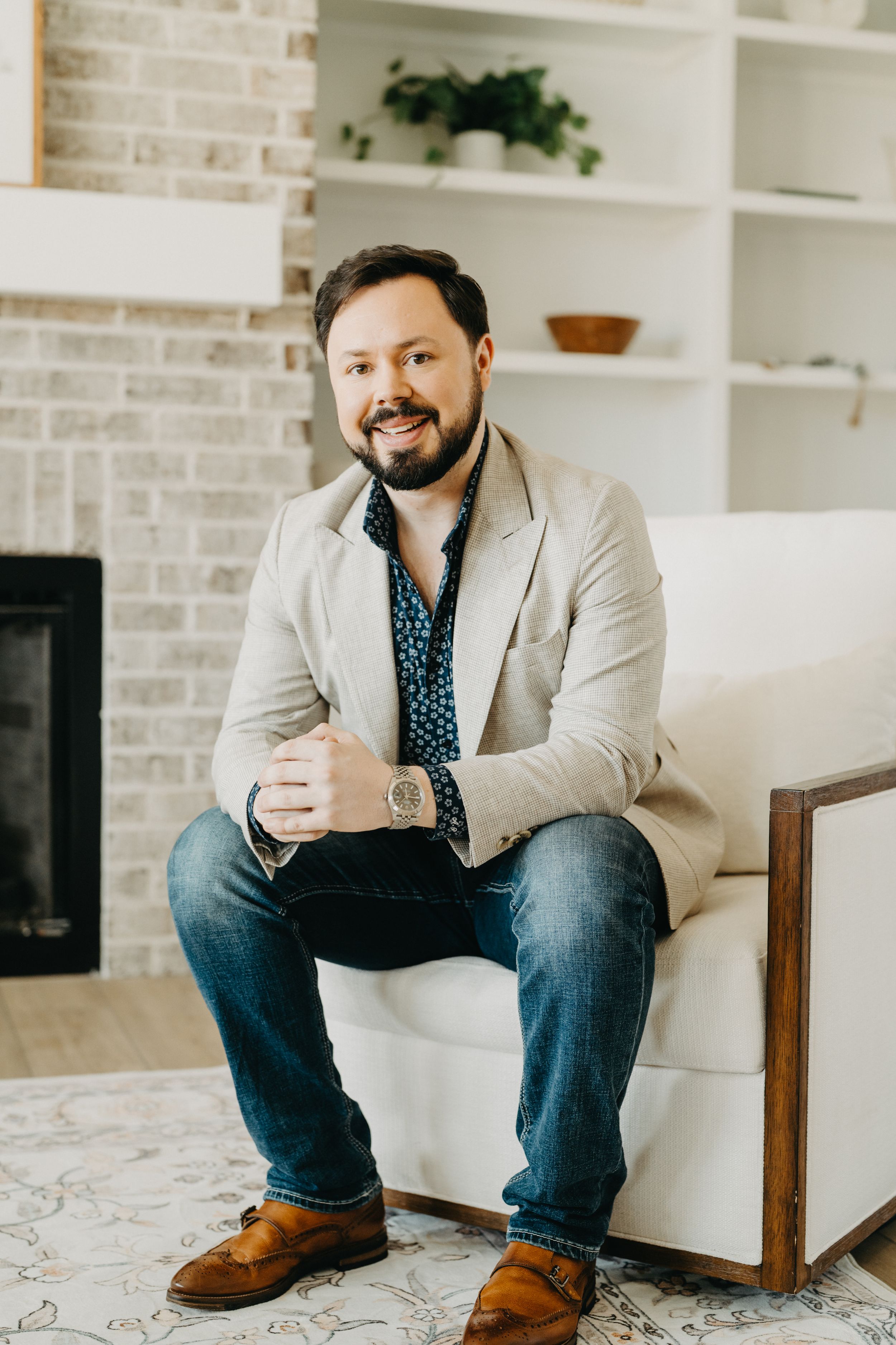 Smiling bearded man in beige blazer and jeans sits relaxed on white chair in modern living room.