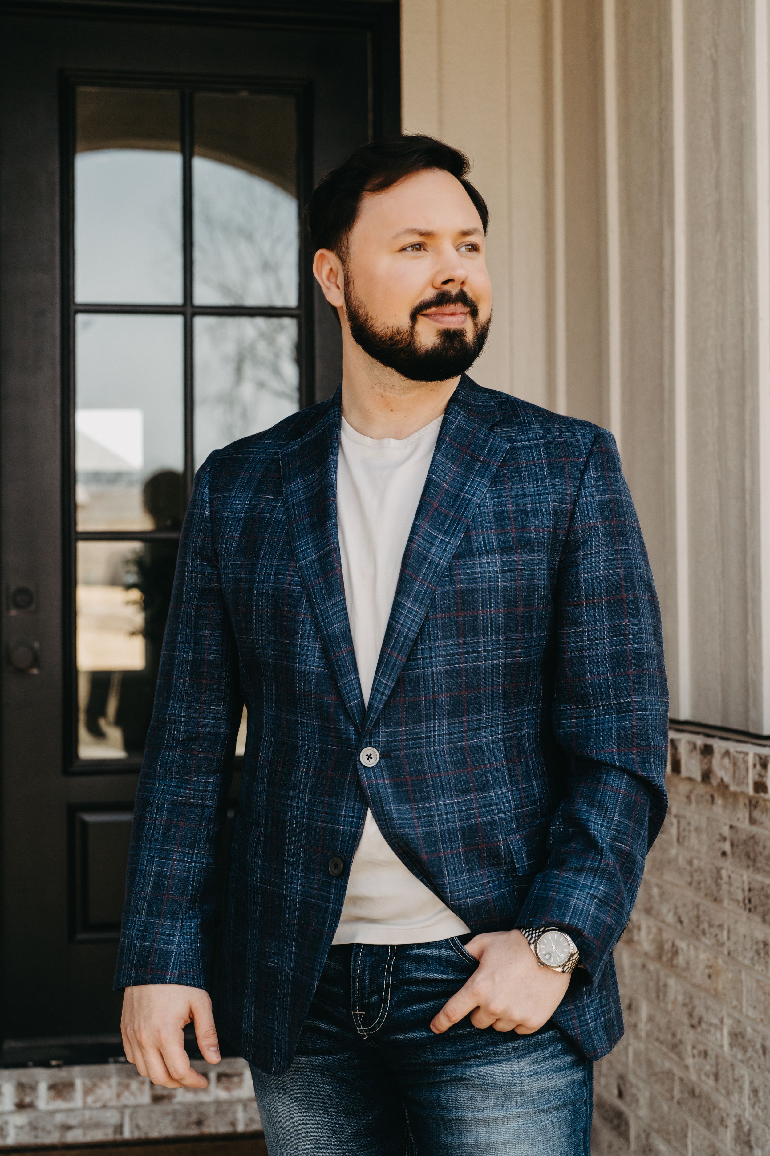 Man in navy plaid blazer and jeans stands confidently by a brick pillar and glass door, looking away thoughtfully.