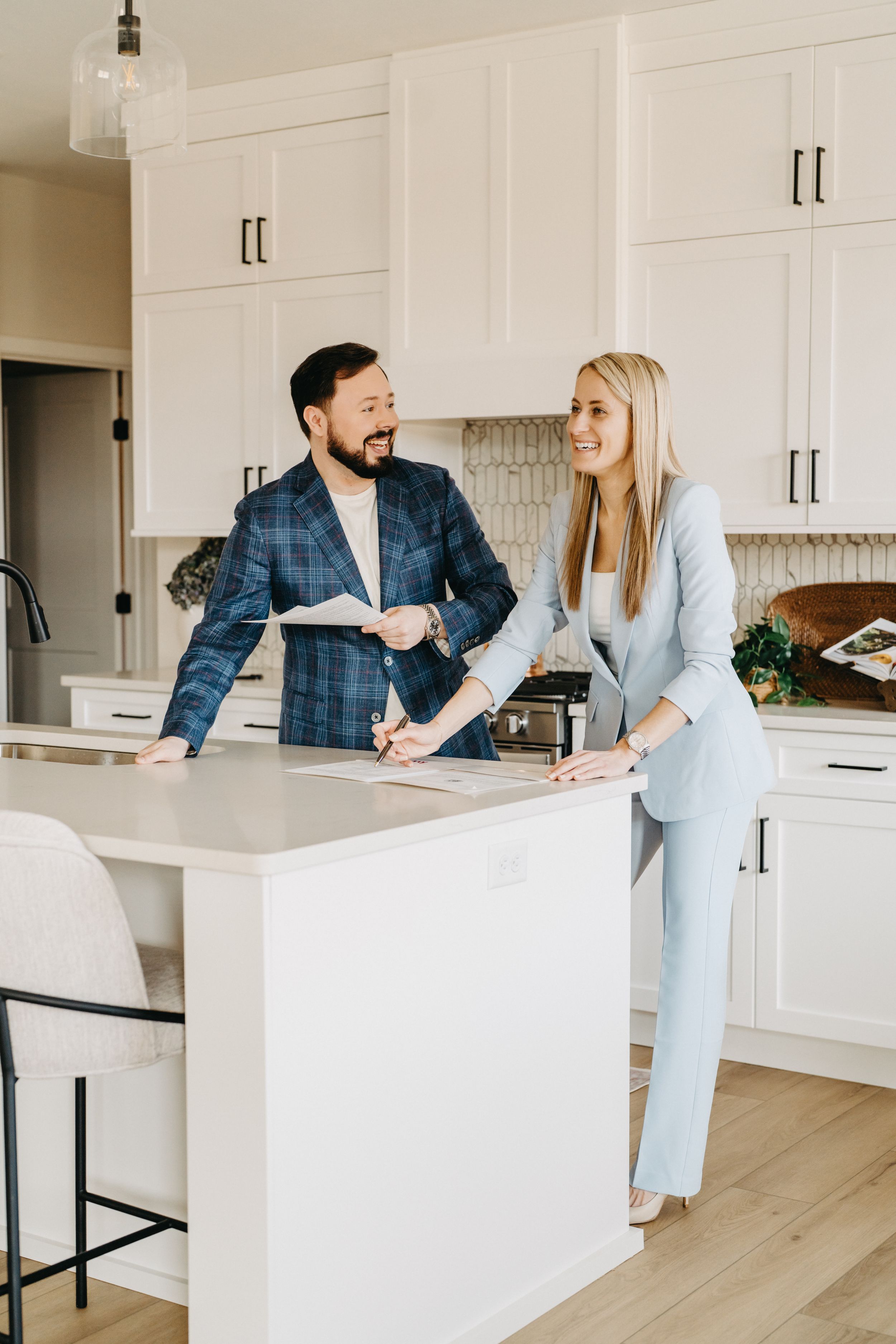 A man and woman in business attire smile while reviewing documents at a white kitchen island.