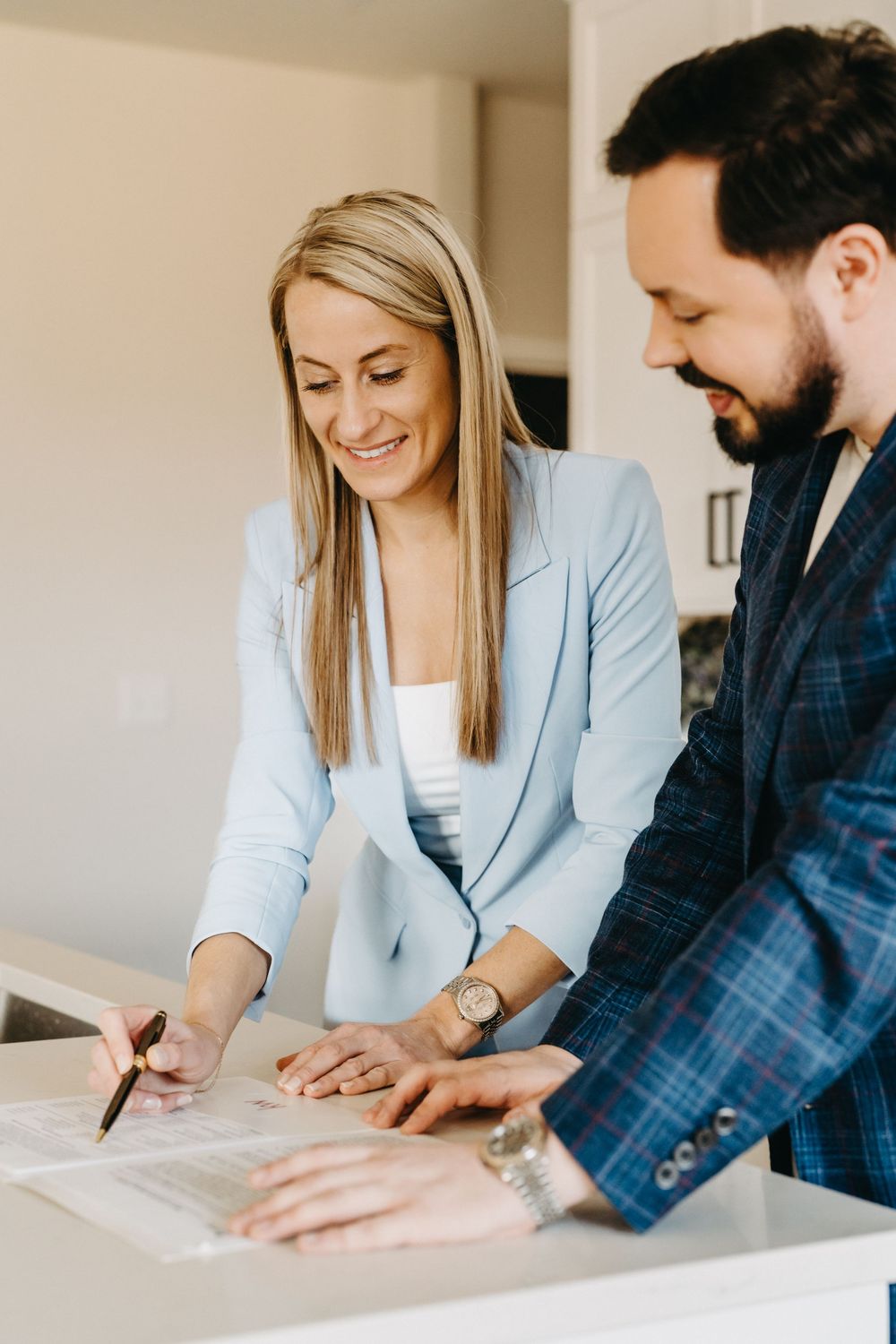 A smiling blonde woman in a blue blazer signs documents with a bearded man in a plaid suit at a white counter.
