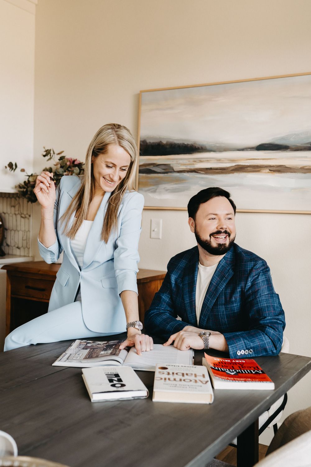 A smiling woman in a light blue suit and a man in a navy blazer review books including Atomic Habits at a wooden table.