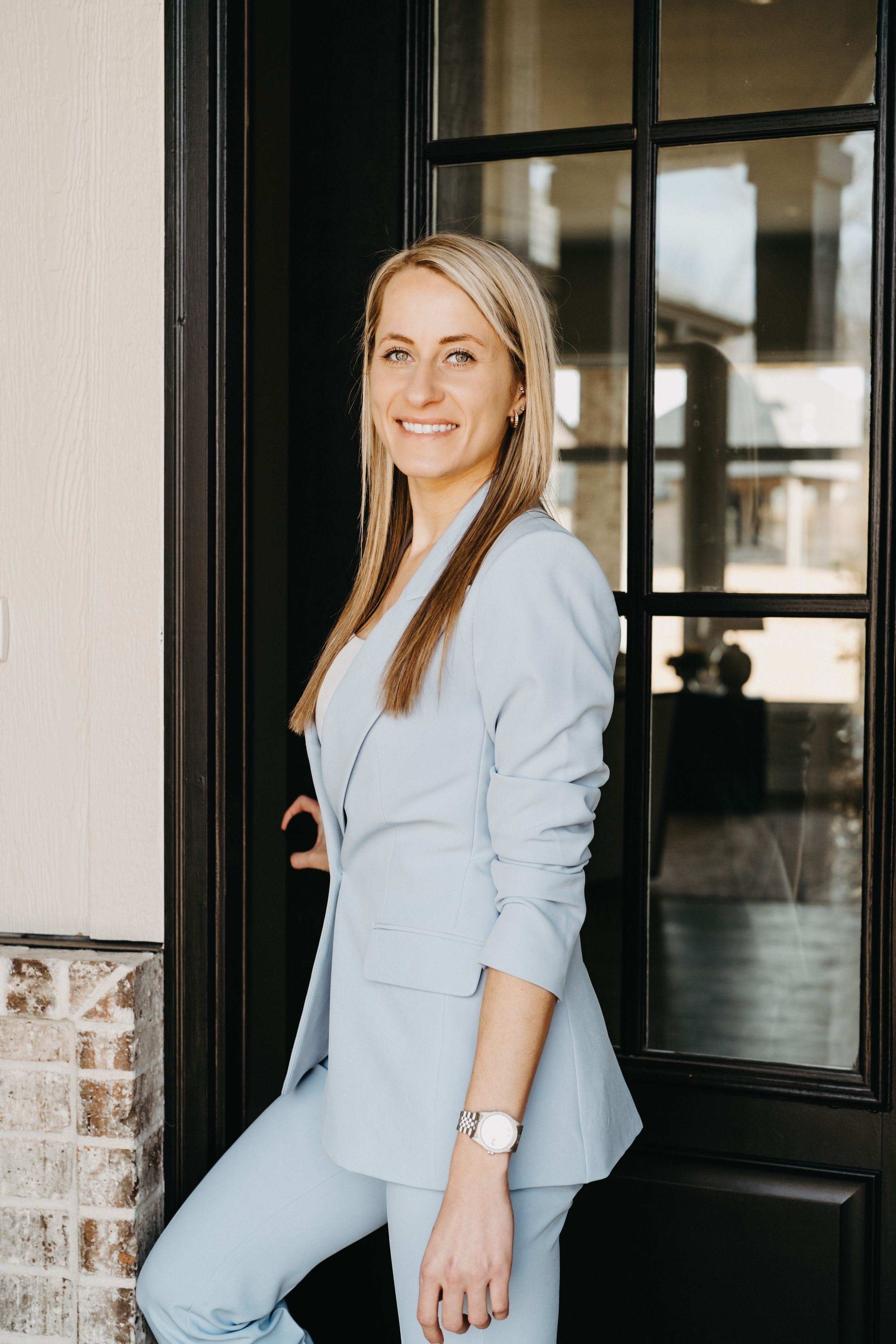 Smiling blonde woman in a light blue blazer and pants suit posing by a black door.