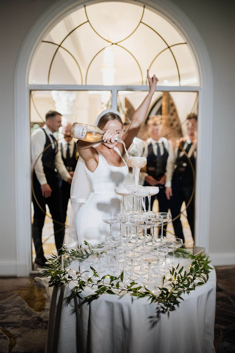 A wedding reception features a champagne tower decorated with greenery on an elegant white-clothed table beneath an arched window.