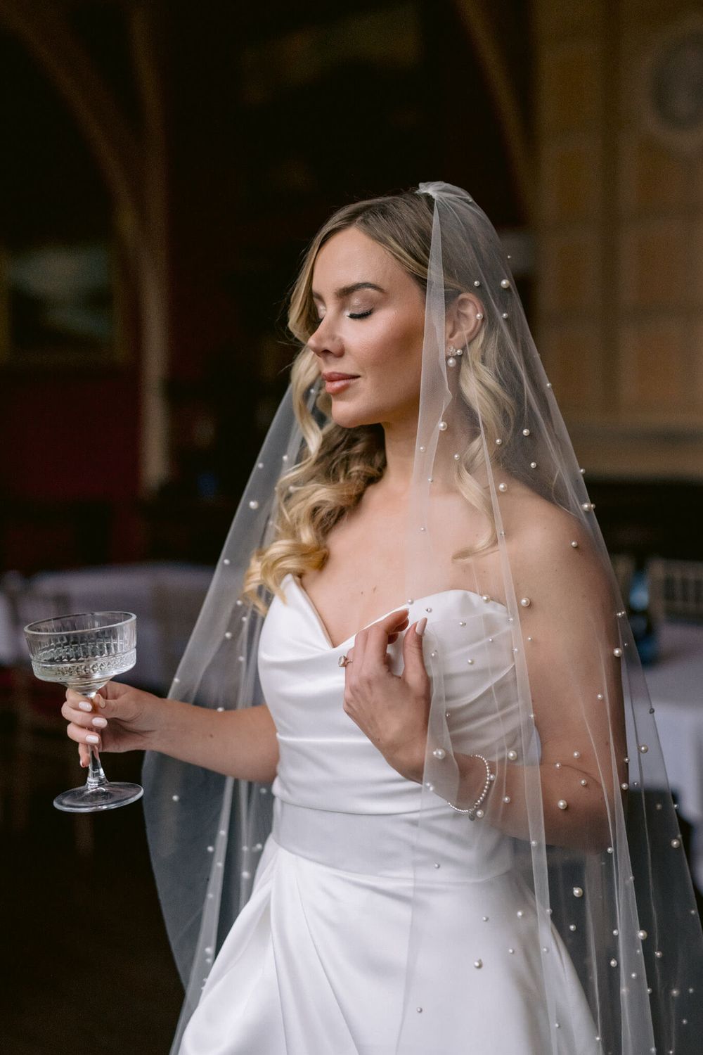 Elegant bride in white gown and pearl-studded veil holding a champagne glass.
