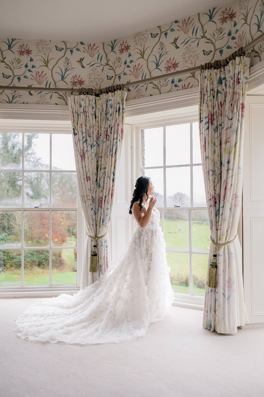 A floral draped bay window setting with an elegant white lace wedding dress train flowing across the light carpeted floor.