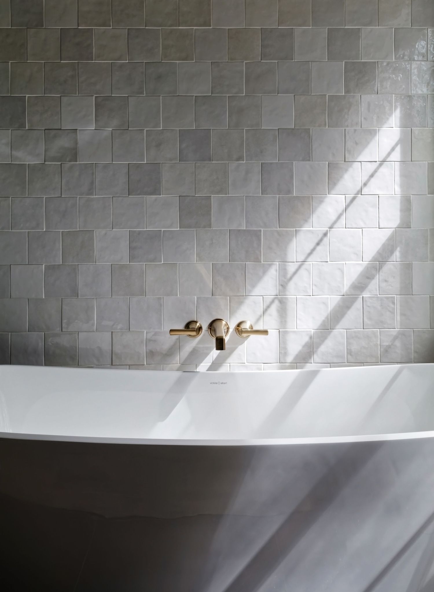Freestanding white soaking tub against grey zellige tile wall with brass wall-mount faucet in natural light.
