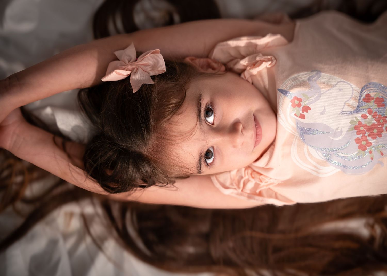 Young girl with hair bow rests on satin fabric in dreamy portrait lighting.