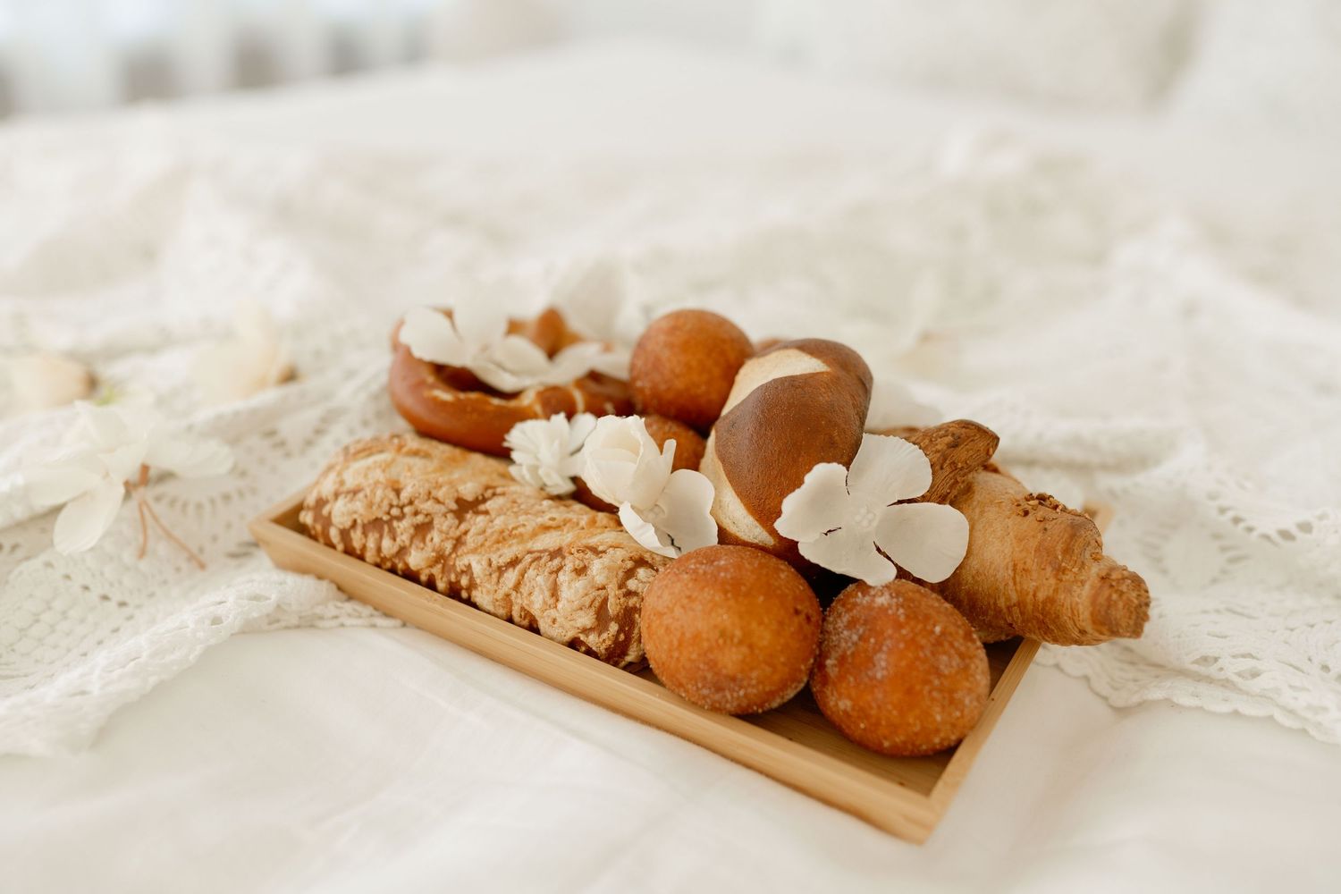 Rustic wooden board with fresh bread rolls and white flowers on lace fabric.