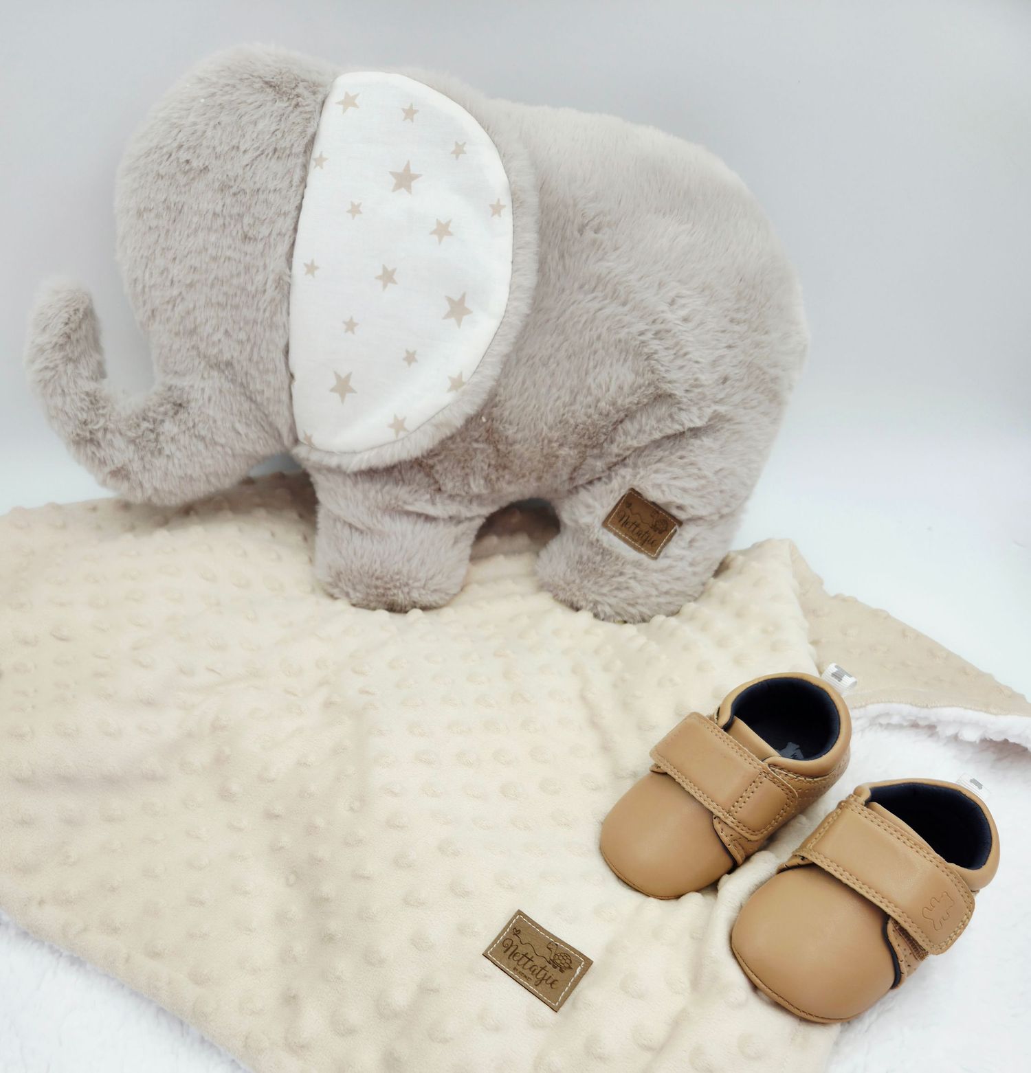 Gray plush elephant with star-patterned ears next to tan leather baby shoes on cream blanket.