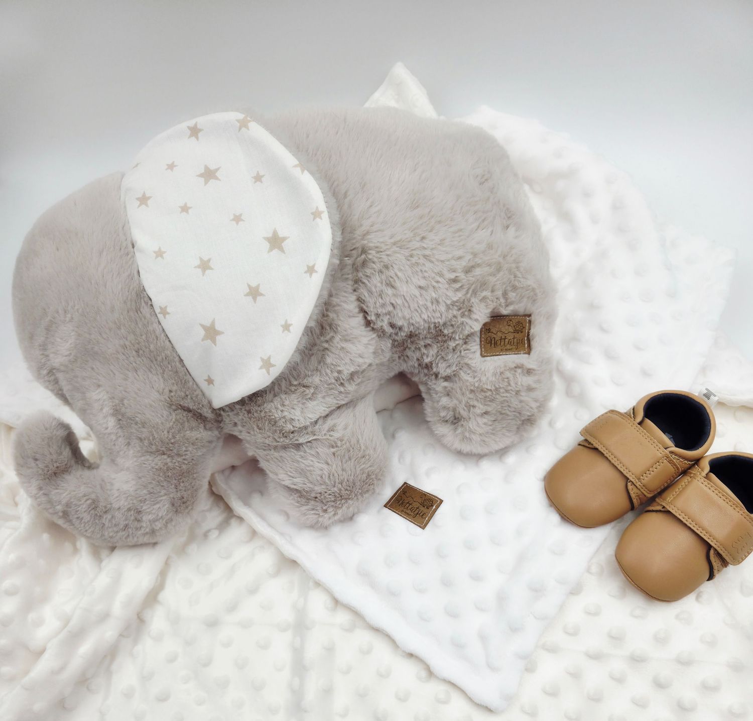 Grey plush elephant with star pattern ears on white textured blanket next to tan shoes.