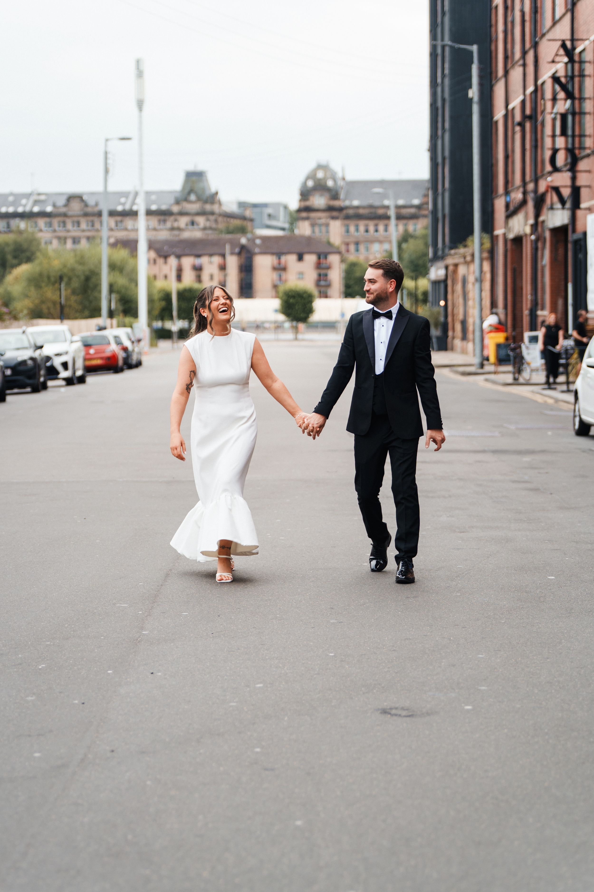 A couple in wedding attire walks and dances playfully together on a city street near industrial buildings.