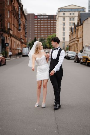 Bride and groom walking hand in hand on a city street, bride in short white dress with veil, groom in black suit.