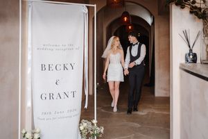 Bride and groom walk hand in hand past a wedding reception sign reading Becky and Grant, 9th June 2024.