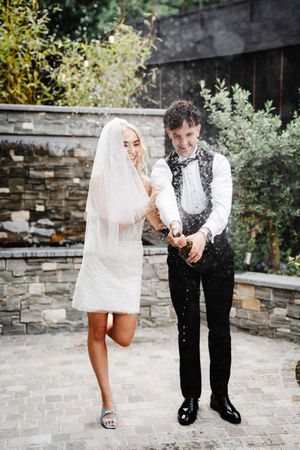 Bride in short white dress and veil laughs with groom as champagne sprays during outdoor wedding celebration.