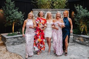 Five women in elegant dresses laughing together outdoors at a garden celebration.