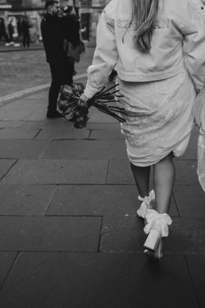 A black and white image shows a person walking on a stone sidewalk wearing a light-colored dress and heels.