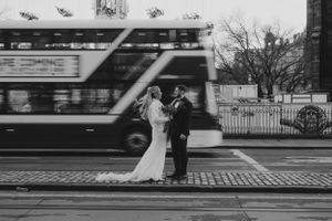 A couple in wedding attire stands on a cobblestone street as a blurred double-decker bus passes by in black and white.