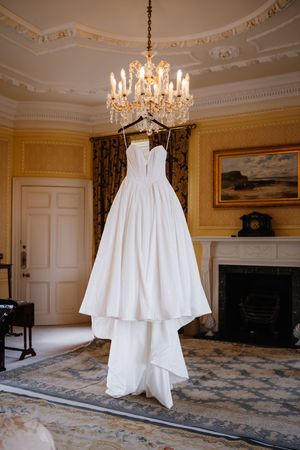Elegant white strapless ball gown wedding dress hanging from a crystal chandelier in a classic yellow-walled room.
