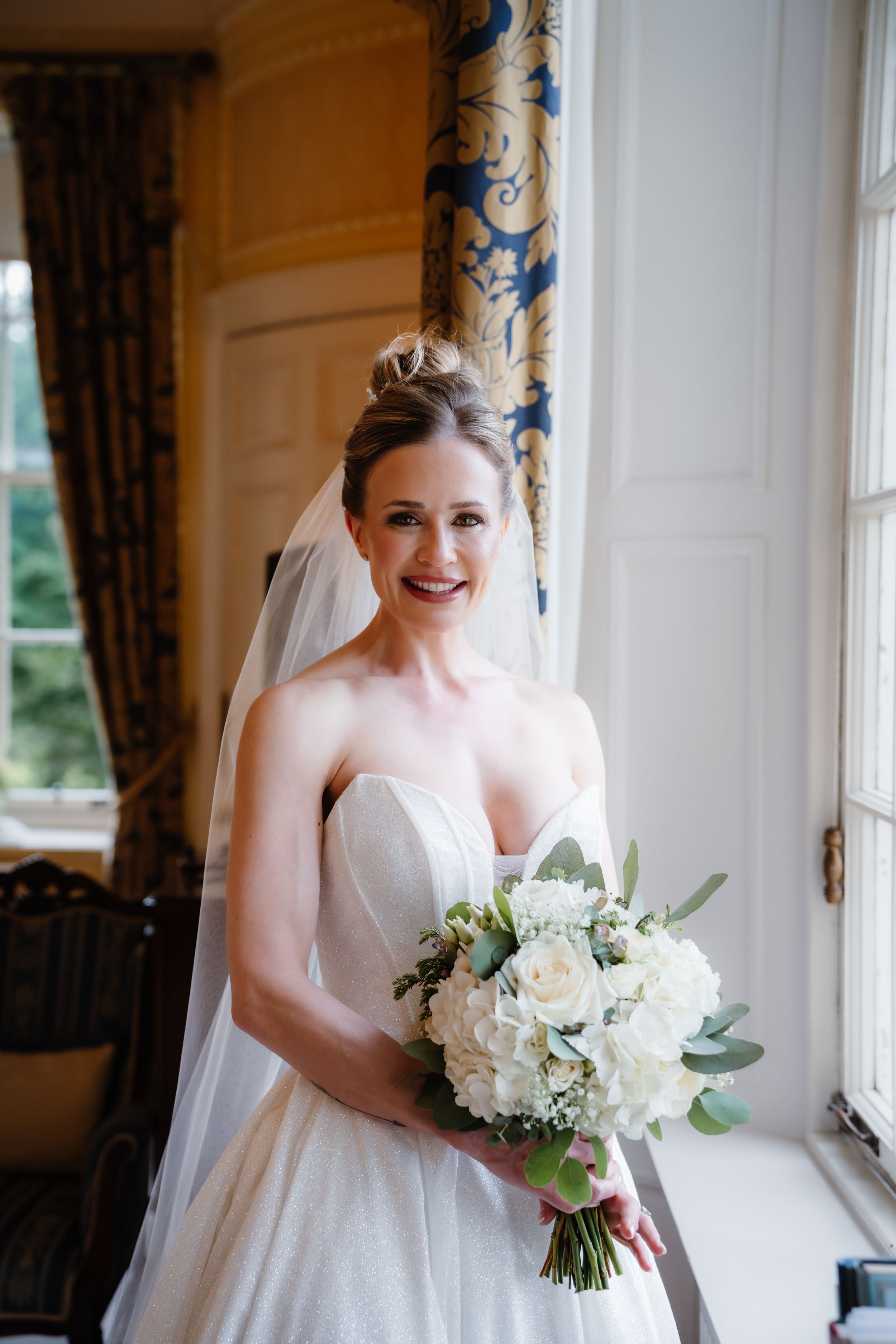 Smiling bride in strapless white gown and veil holds white hydrangea bouquet by elegant window.