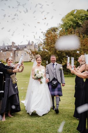Bride and groom walk through confetti at Scottish castle wedding, groom wearing traditional tartan kilt.