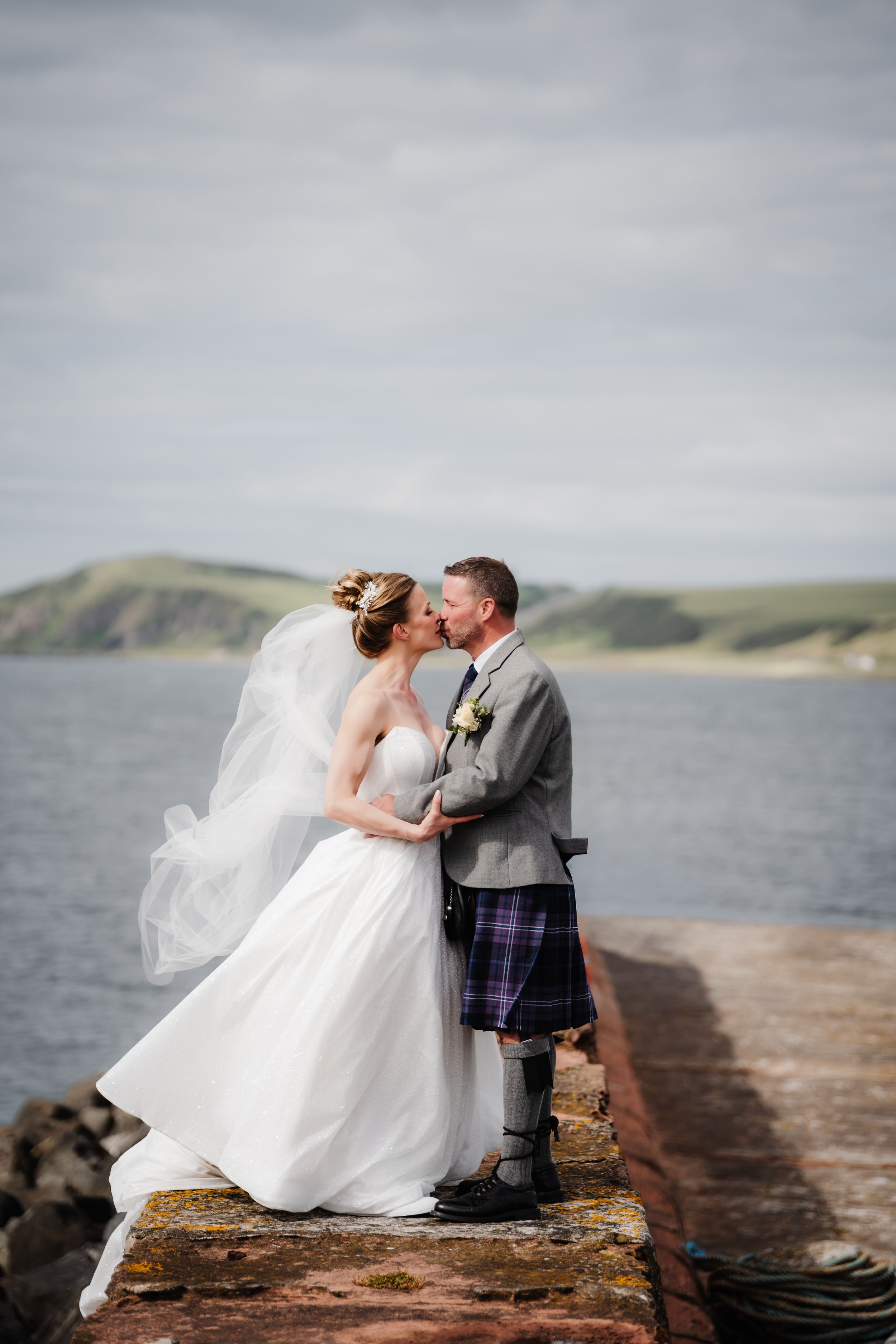Bride and groom kissing on a seaside pier, groom wearing a traditional Scottish kilt, with veil blowing in the wind.