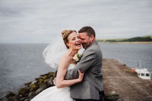 Joyful bride and groom embracing on a seaside pier, with veil blowing in the wind on a cloudy day.