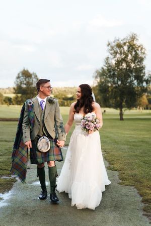 A couple in wedding attire walks together on a grassy field, with the groom wearing a traditional Scottish kilt and jacket.
