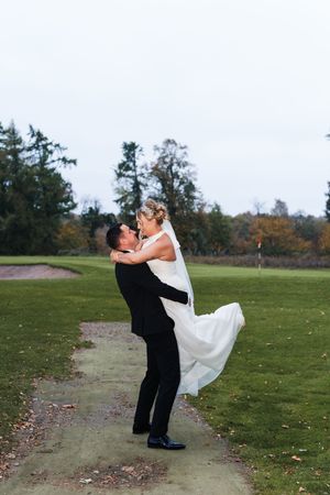 A couple in wedding attire playfully dips and poses together on a grassy field during an autumn day.