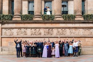 Wedding party and guests line up for a panoramic photo in front of a grand stone building with columns.