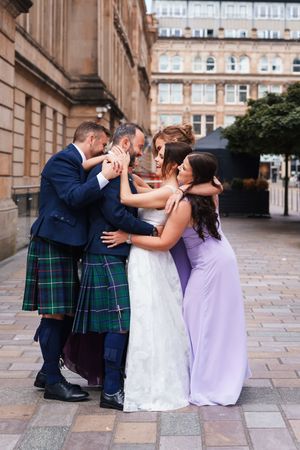 Wedding party members in kilts and formal attire share joyful moments on stone pavement.