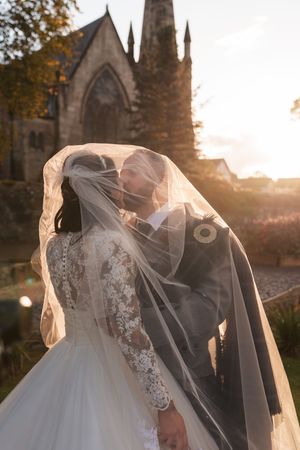 A romantic wedding couple embraces under a flowing veil at sunset with a gothic church spire in the background.