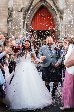 Wedding guests throw confetti at a couple outside a church as the bride's white dress swirls in celebration.